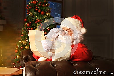 Portrait of happy Santa Claus sitting at his room at home near Christmas tree and reading Christmas letter or wish list. Stock Photo