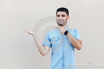 Portrait of happy satisfied handsome young bearded man in blue shirt standing, holding, pointing something and looking at camera Stock Photo