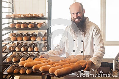 Portrait of happy smiling proud young adult baker with long beard in white uniform standing in his workplace and holding full tray Stock Photo