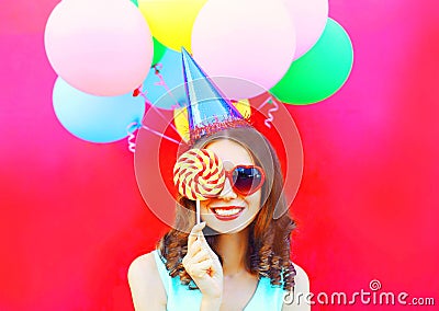 Portrait happy smiling woman in a birthday cap closes her eye with a lollipop on stick over an air colorful balloons on pink bac Stock Photo