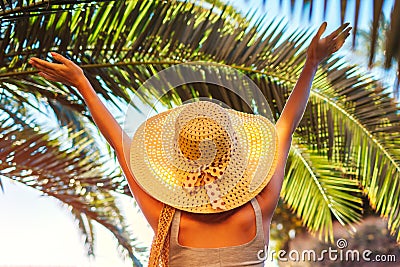 Portrait of happy woman in hat lifting hands under palm leaf in hotel yard. Summer vacation Stock Photo