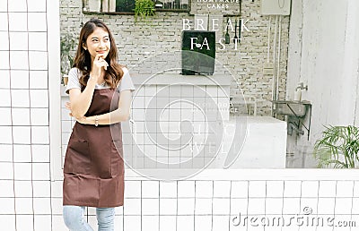Portrait of a happy young Asian barista in apron looking away and thinking in front of her small coffee shop, waist up, copy space Stock Photo