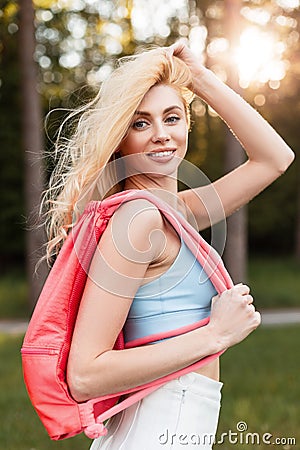 Portrait happy young blonde woman with cute smile in fashionable blue top with pink sports backpack in white skirt in forest at Stock Photo