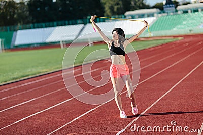 Portrait of happy young sports brunette woman in black top and rose shorts outdoors on stadium holding skipping rope over the head Stock Photo