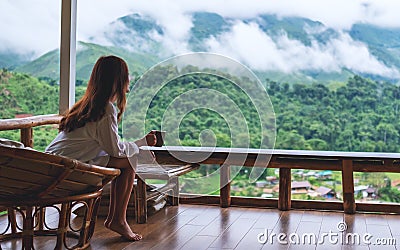 A beautiful asian woman holding and drinking hot coffee , sitting on balcony and looking at mountains and green Stock Photo