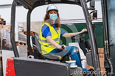 Portrait industrial female worker wearing a medical mask using a forklift in a warehouse wearing a yellow vest Stock Photo