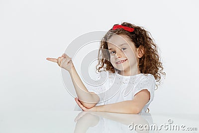 Portrait of a little adorable curly girl, seated at table, showing with finger on direction, over white background. Stock Photo