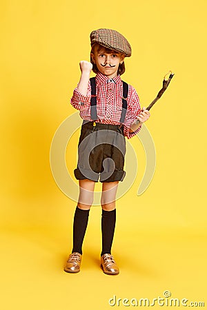 Portrait of little artisitc girl, child in retro shirt and shorts, holding slingshot against yellow studio background Stock Photo