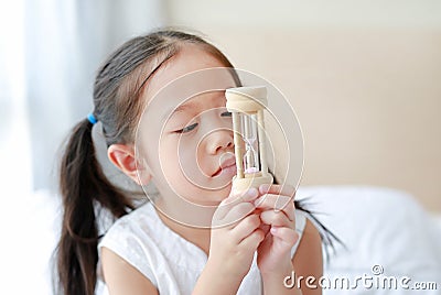 Portrait of little Asian girl looking at hourglass in hand lying on bed at home. Waiting times with sandglass Stock Photo