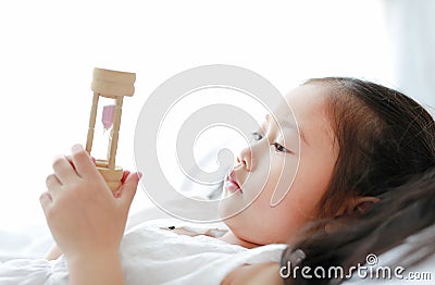 Portrait of little Asian girl looking at hourglass in hand lying on bed at home. Waiting times with sandglass. Close-up shot Stock Photo