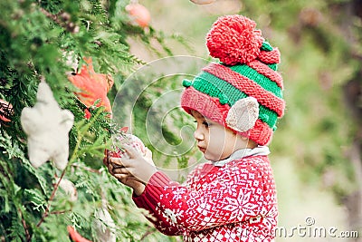 Portrait of a little boy in elf hat and red sweater near the christmas tree and holding decoration Stock Photo