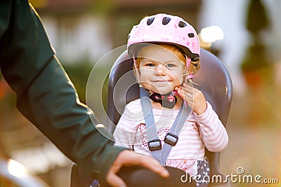 Portrait of little toddler girl with security helmet on the head sitting in bike seat and her father or mother with Stock Photo
