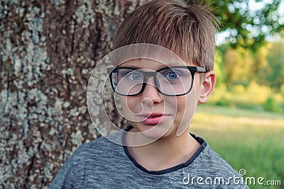 Loveable boy with blue eyes, in glasses in gray longsleeve standing in front of tree in park and enjoying fresh air. Stock Photo