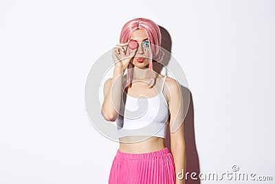 Portrait of lovely girl in pink wig, dressed up for party or holiday celebration, holding delicious macaroon and pouting Stock Photo