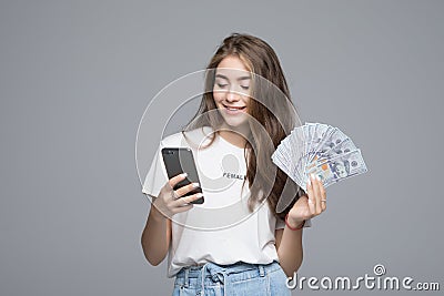 Portrait of lucky young woman smiling while holding fan of money and cell phone in hands over gray background Stock Photo