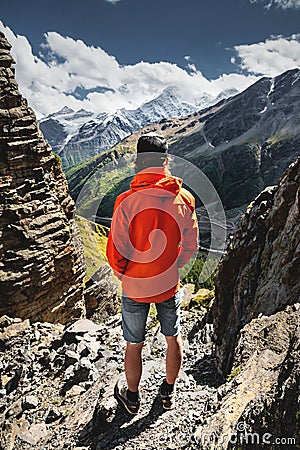 Portrait of a male hiker in shorts and a cap from the back is standing on high rocks at the edge of a cliff against the Stock Photo