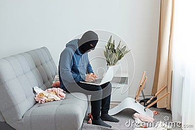 Portrait of man wearing blue hoodie and black robber mask, breaking flat, sitting on sofa and opening laptop, stealing modern Stock Photo