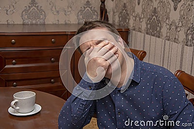 Portrait of a man 30-35 years old, who covered his face with his hands while sitting at a table. Perhaps he is upset or sad, cryin Stock Photo