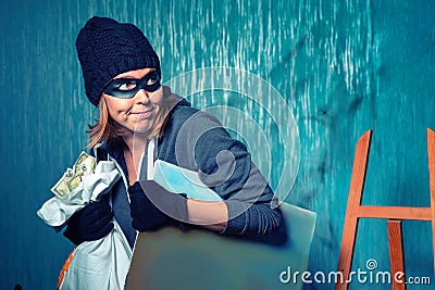 Portrait of a masked female robber is carrying a stolen bag of money and a painting. The background is dark blue. The concept of Stock Photo