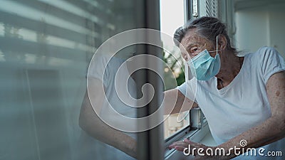 Portrait of masked senior woman in late 90s waving from home window. Elderly woman in protective mask looks out the Stock Photo