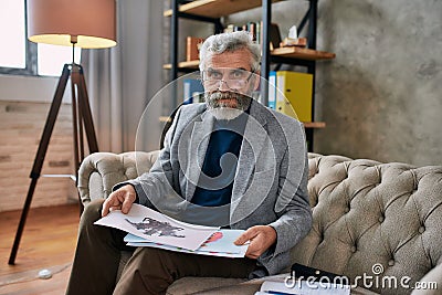 Portrait of mature psychologist in glasses looking at camera, holding pictures with ink stain, Rorschach Inkblot sitting Stock Photo