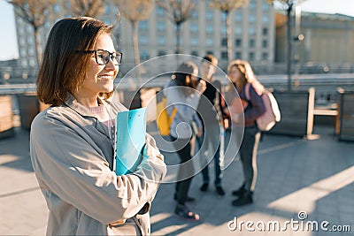 Portrait of mature smiling female teacher in glasses with clipboard, outdor with a group of teenagers students, golden hour Stock Photo