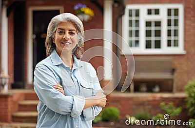 Portrait Of Mature Woman Standing In Garden In Front Of Dream Home In Countryside Stock Photo