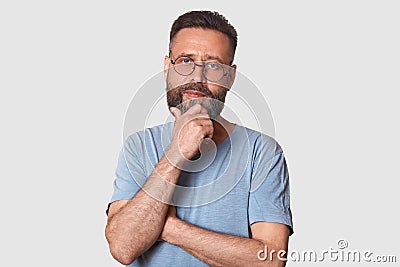 Portrait of middle bearded aged male with pensive facial expression, dressed gray cassual t shirt and round spectacles, keeps hand Stock Photo