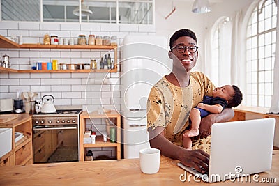 Portrait Of Multi-Tasking Father Holding Sleeping Baby Son And Working On Laptop Computer In Kitchen Stock Photo