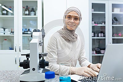 Portrait of Muslim young woman in hijab, chemist scientist working in laboratory at laptop and writing information and Stock Photo