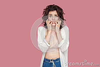 Portrait of nervous worried brunette young woman with curly hairstyle in casual style standing, confused face, bitting her nails Stock Photo