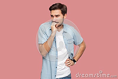 Portrait of nervous worry handsome bearded young man in blue casual style shirt standing looking away and bitting his nails Stock Photo