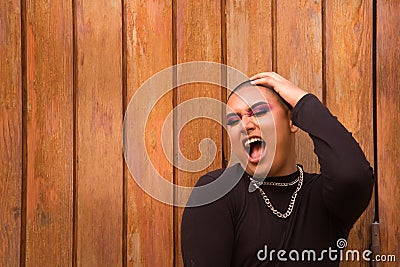 Portrait of non-binary person, young and South American, heavily make up and with one hand on head screaming with a wooden wall in Stock Photo