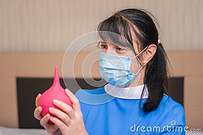 Portrait of a nurse in a medical mask with a red enema in hands. A woman in medical uniform is preparing to do a wash to the Stock Photo