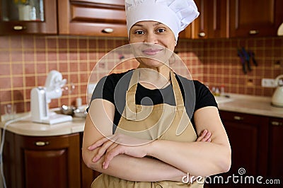 Pleasant housewife in chef's hat and apron, standing with arms crossed in the home kitchen, smiling, looking at Stock Photo