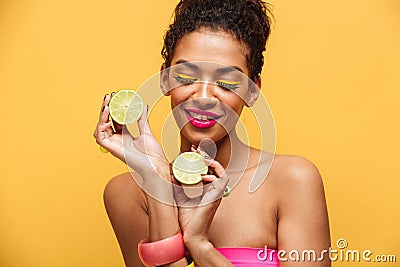 Portrait of pleased afro american woman with trendy makeup holding two halves of fresh lime in both hands isolated, over yellow b Stock Photo