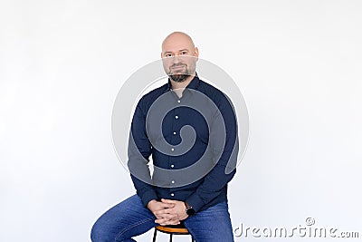 Portrait of pleased middle-aged man wear blue shirt, jeans sitting on stool, interlacing fingers on white background. Stock Photo