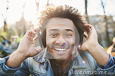 Portrait of positive adult dark-skinned man smiling broadly while sitting in park, listening to music in headphones and Stock Photo