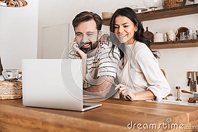 Portrait of positive couple looking at laptop while cooking pastry in kitchen at home Stock Photo