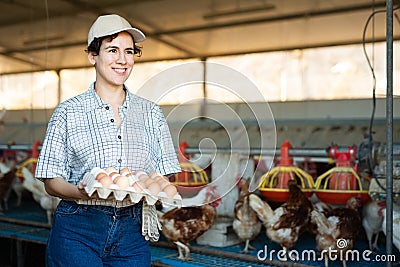 Smiling adult latin woman in plaid shirt and cap collecting eggs in chicken Stock Photo