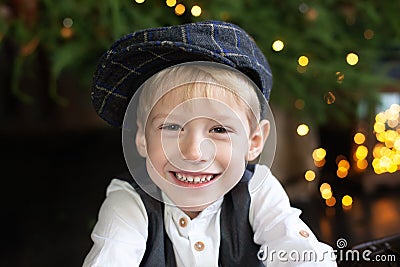 Portrait of positive little boy in cap against backdrop of Christmas tree with garland lights. Sincere child emotions. Happy child Stock Photo