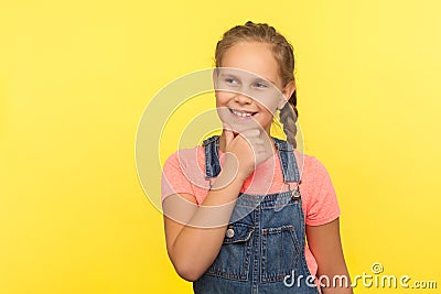 Portrait of positive thoughtful little girl in denim overalls holding chin and dreaming with cunning expression Stock Photo