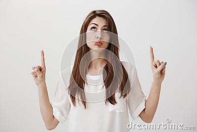 Portrait of pouting gloomy caucasian girl pointing and looking up with regret, standing over gray wall, being childish Stock Photo