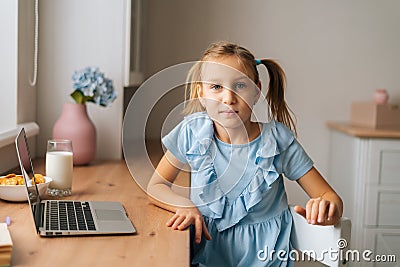 Portrait of pretty elementary child girl sitting at home table with laptop and snack by window, looking at camera. Editorial Stock Photo