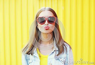 Portrait pretty young woman in red sunglasses blowing lips kiss over yellow Stock Photo