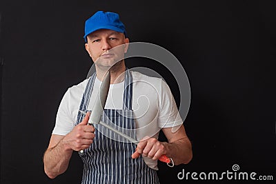 Portrait of a professional butcher in black and white apron, white t shirt and blue baseball hat. Sharpening his knives on a steel Stock Photo