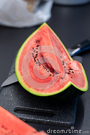 A portrait of a quarter slice of a green cut watermelon lying on a plastic cutting board next to a knife. The piece of the fruit Stock Photo