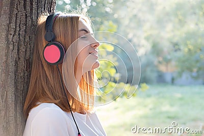 Portrait of red haired teenage girl listening to music in big pink earphones outdoors leaning to a tree Stock Photo