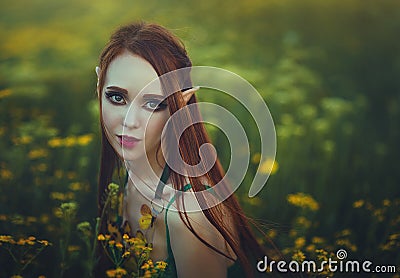 Portrait of a redheaded girl elf in a green swimsuit posing in a clearing of yellow flowers. Fantastic young woman with Stock Photo