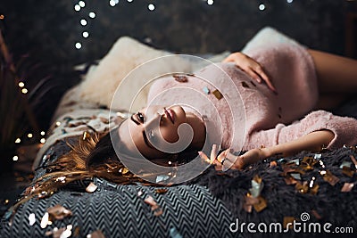 Portrait of sad attractive young woman with tinsel confetti and garland lights celebrating alonein dark room. New year`s Stock Photo